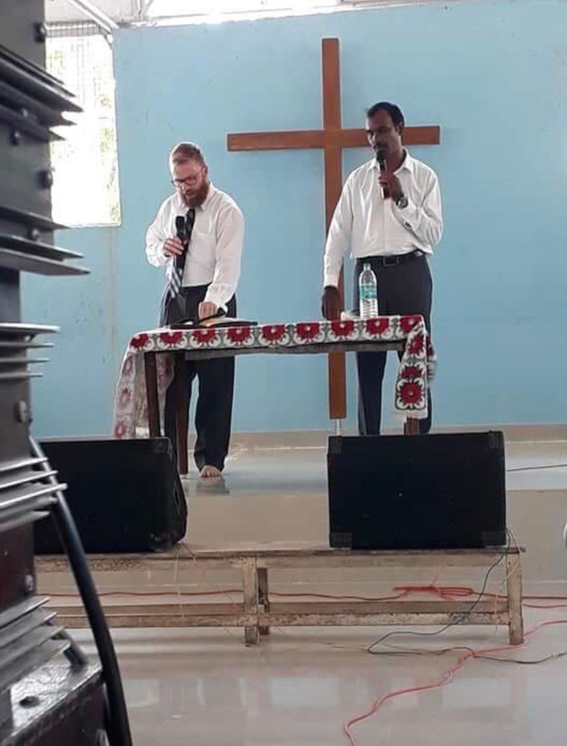 two men are standing at a table with microphones in front of a cross .