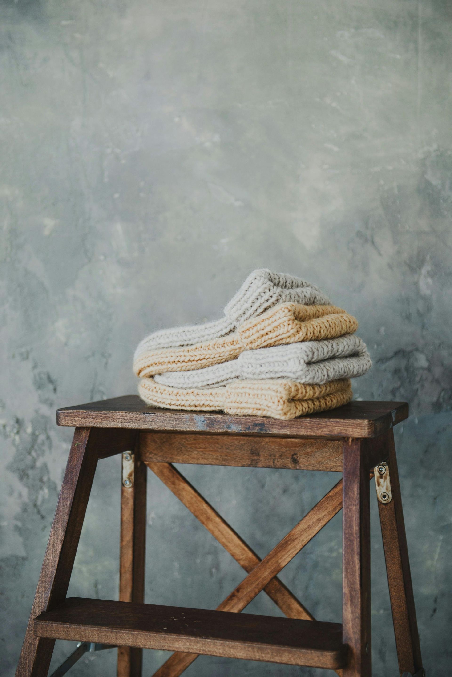 Stacked knitted socks, light brown and grey, on a wooden step stool against a blue-gray wall.