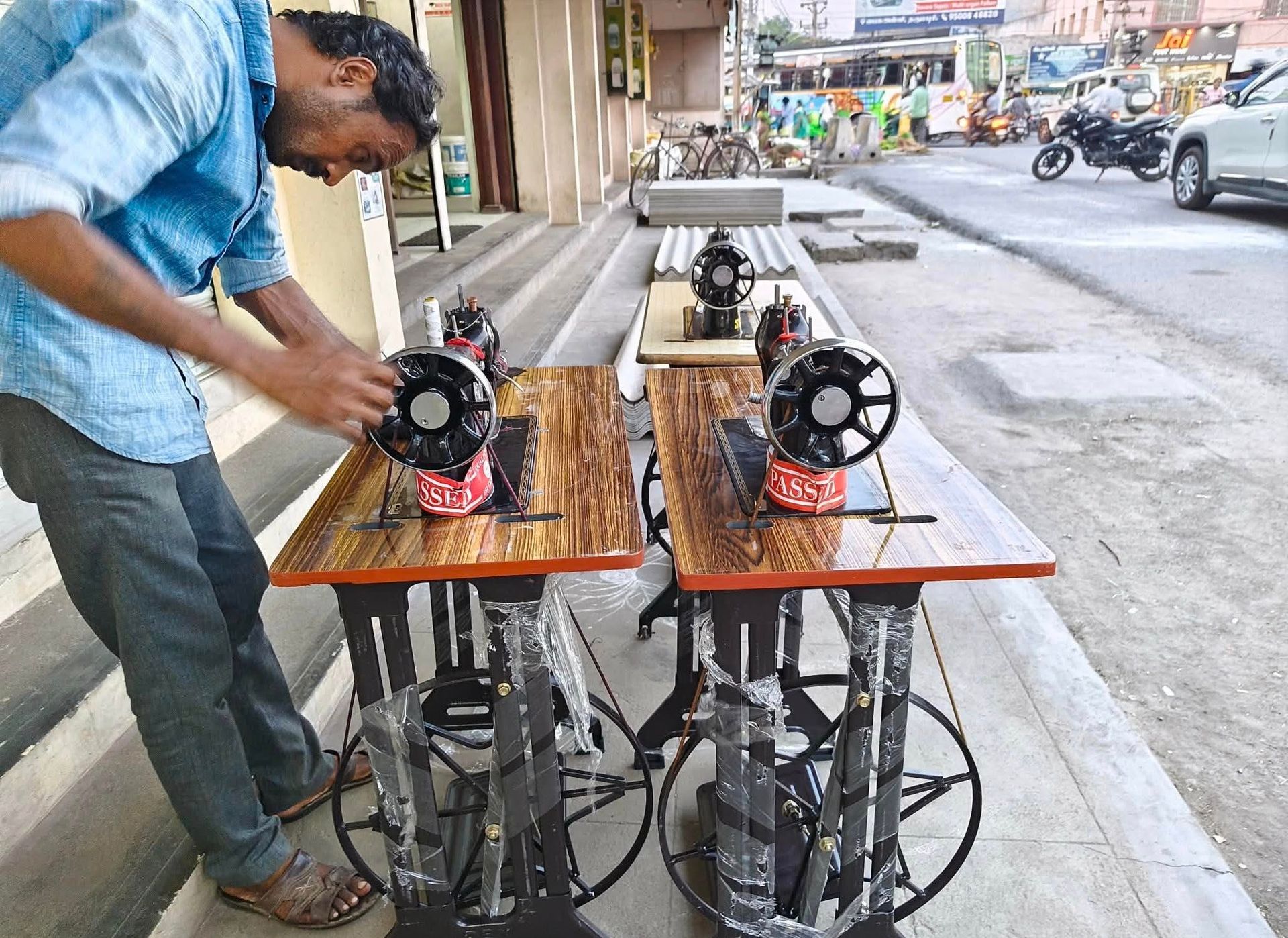 A person works on one of two vintage-style sewing machines set up on a sidewalk along a street.