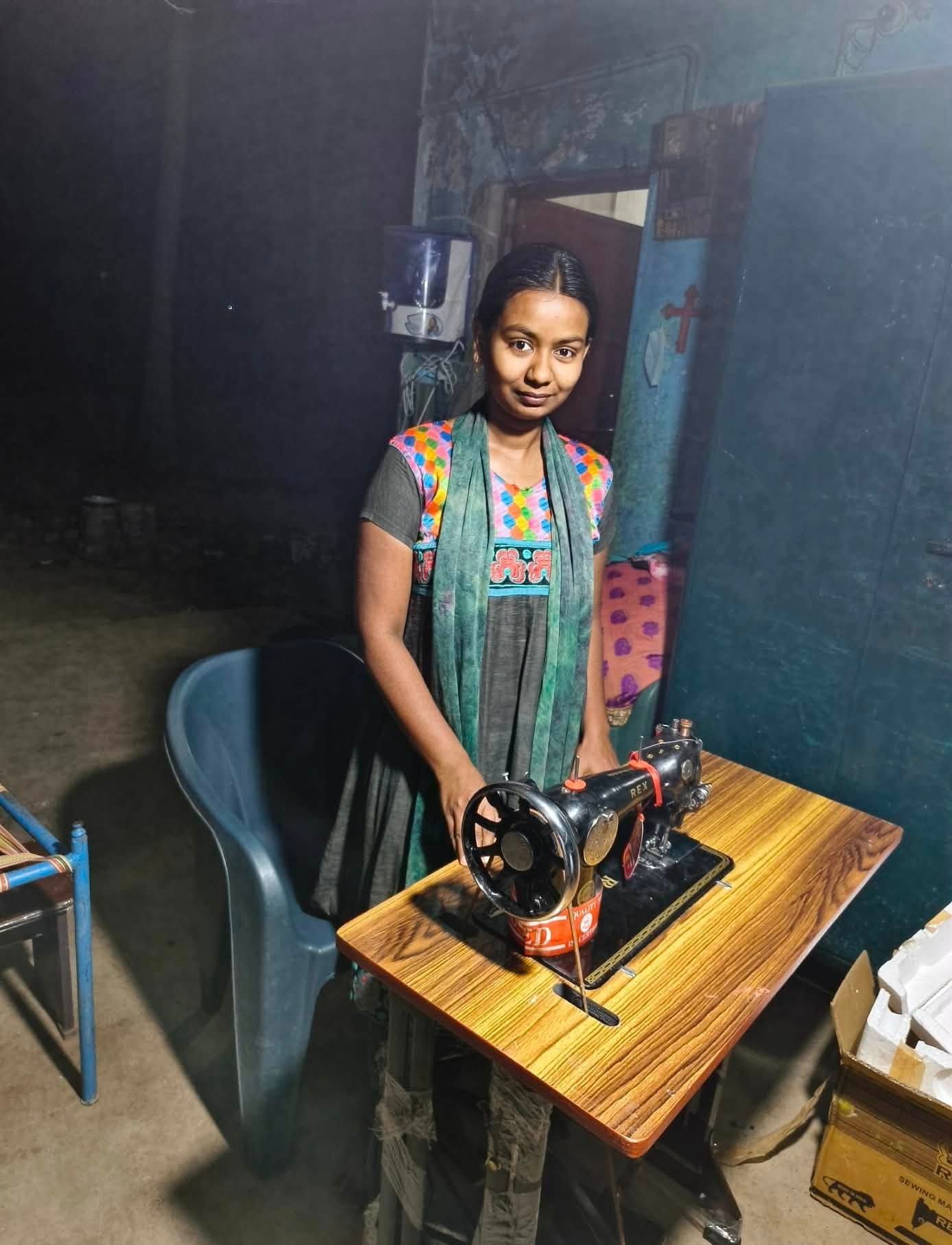 A person wearing a patterned top and teal scarf stands at a wooden table operating a vintage black sewing machine.