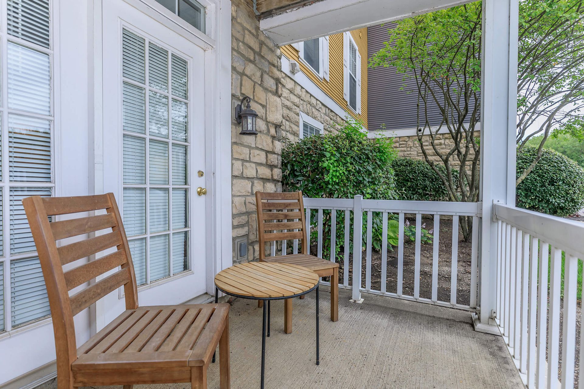 Wooden chairs and table on a porch, near a door. Brick wall and white railing visible.