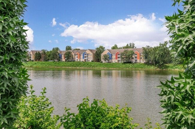 Apartment buildings with colorful roofs by a lake, framed by green trees against a cloudy blue sky at Quarry in Columbus, OH.