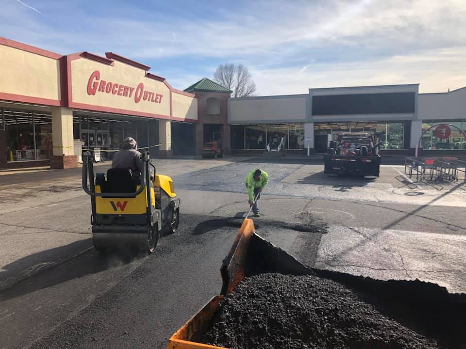 A man is driving a roller on a road in front of a grocery store.