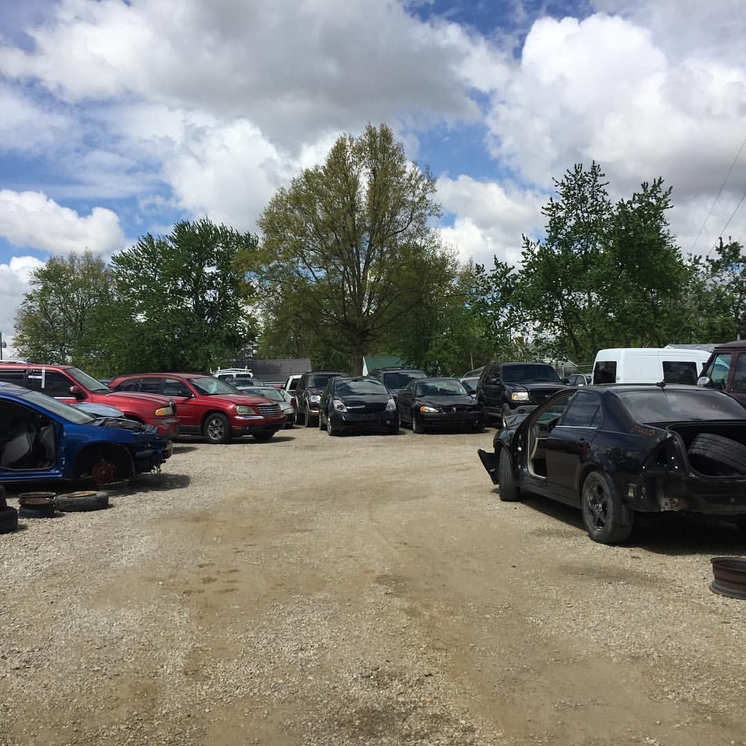 Junk yard with various vehicles, under cloudy skies. Some cars are missing parts.