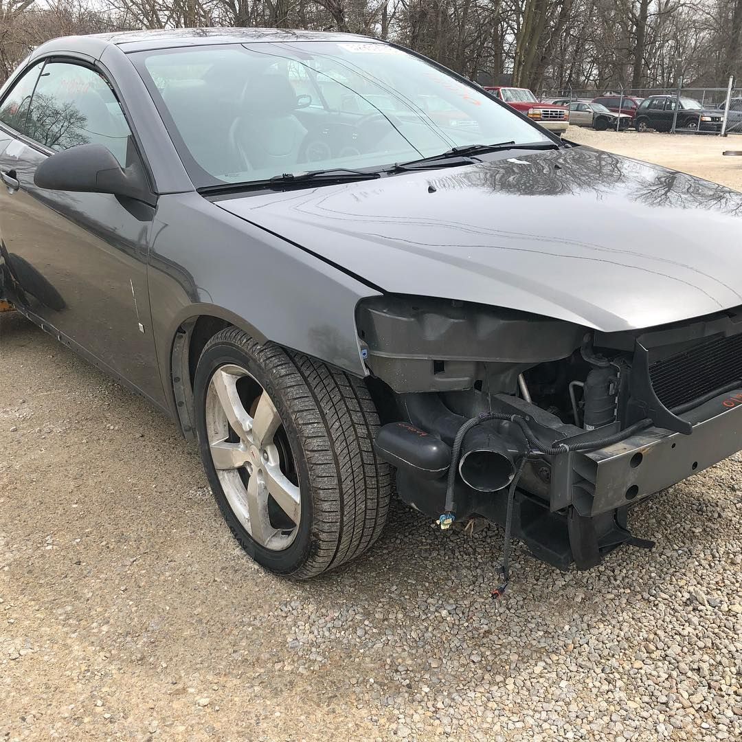 Damaged gray car missing its front bumper, parked on gravel lot.