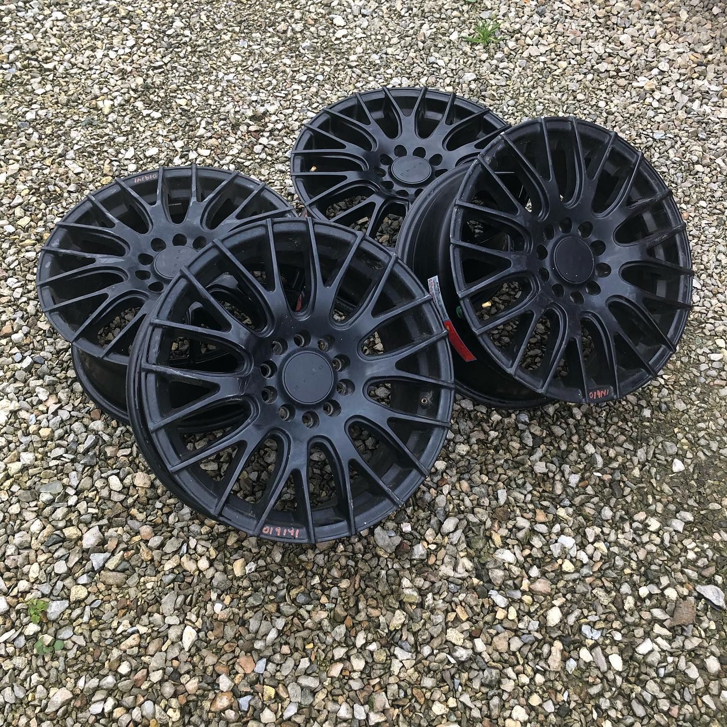 Four black alloy car wheels on a gravel surface.