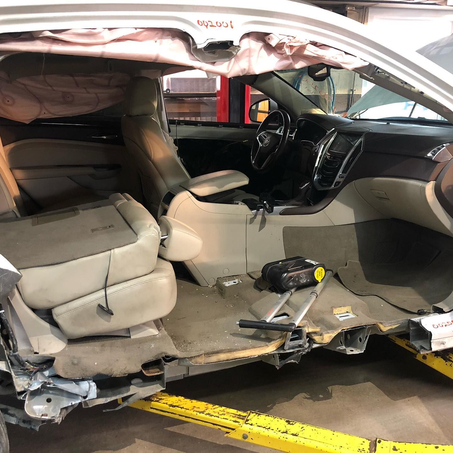 Interior view of a damaged white SUV with deployed airbags. Shows the driver's side and dashboard, on a lift.