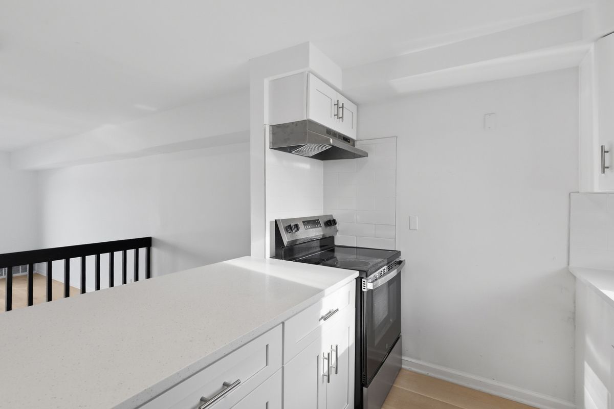 White kitchen with countertop, cabinets, stove, and range hood. A dark railing is visible in the background.