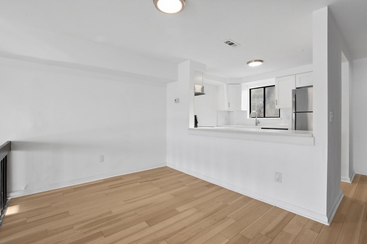 Empty apartment interior with light wood floors, white walls, and a small kitchen visible in the background.