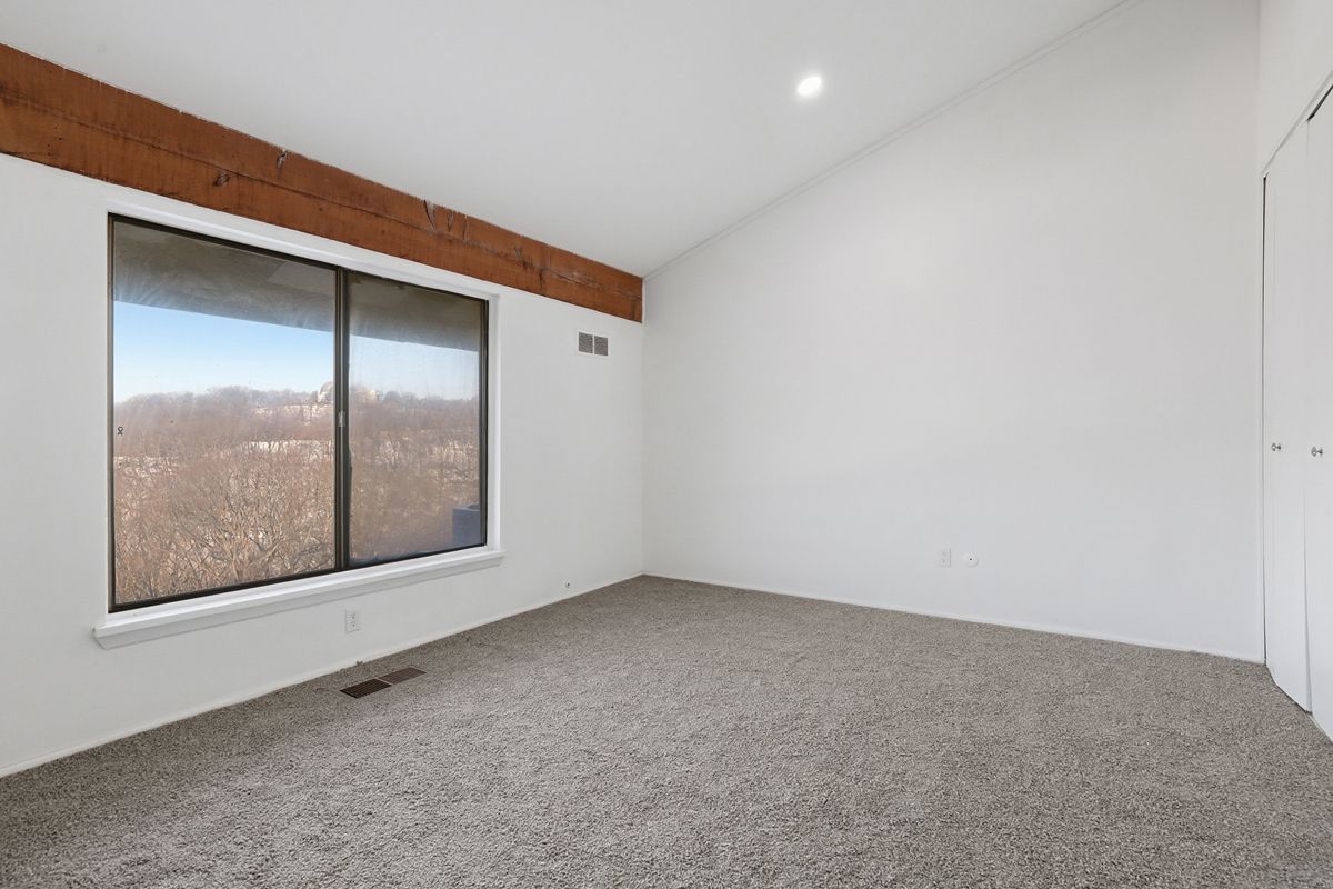 Empty bedroom with a large window and brown wood beam, carpeted floor, and white walls.