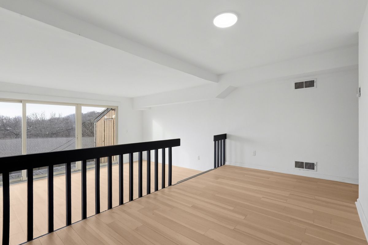 Loft interior with black railing, wooden floor, and large window overlooking trees. Bright, white walls and ceiling.