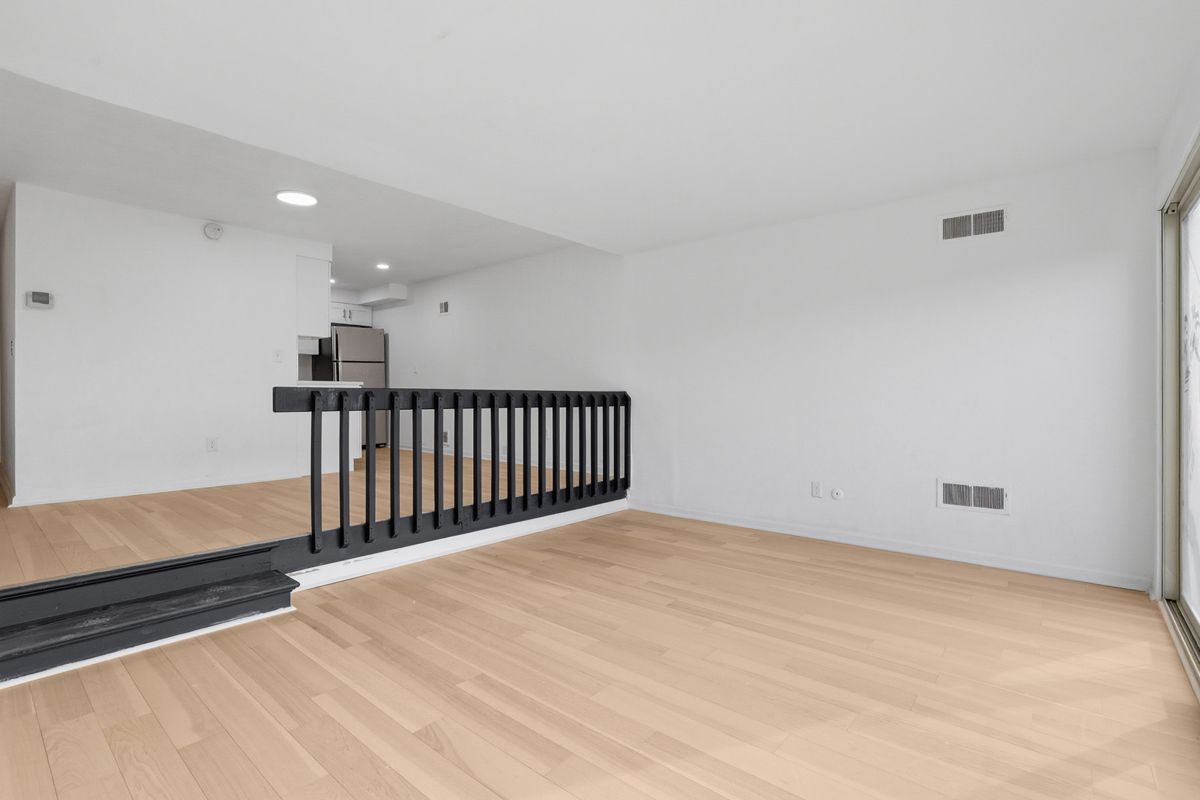 Empty room with light wood floors, white walls, black railing, and a view into a small kitchen.
