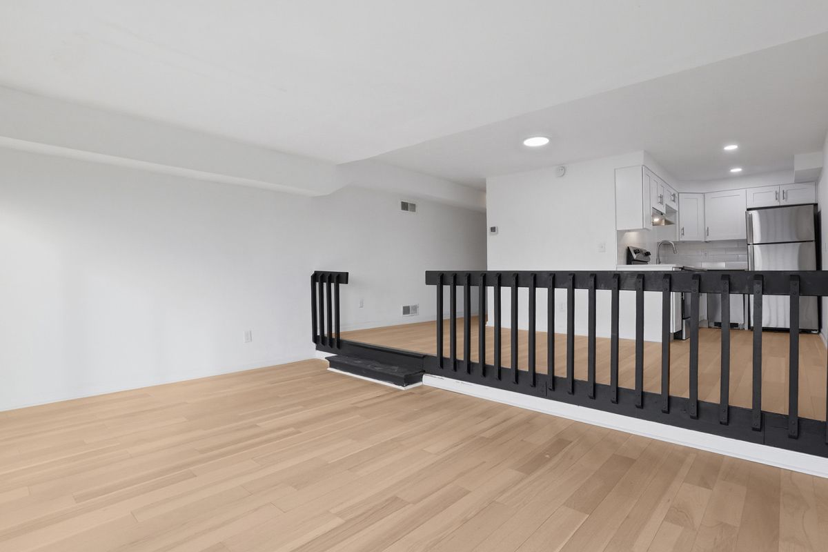 Open-plan living space with light wood floors, a kitchen, and a dark railing on a raised area.