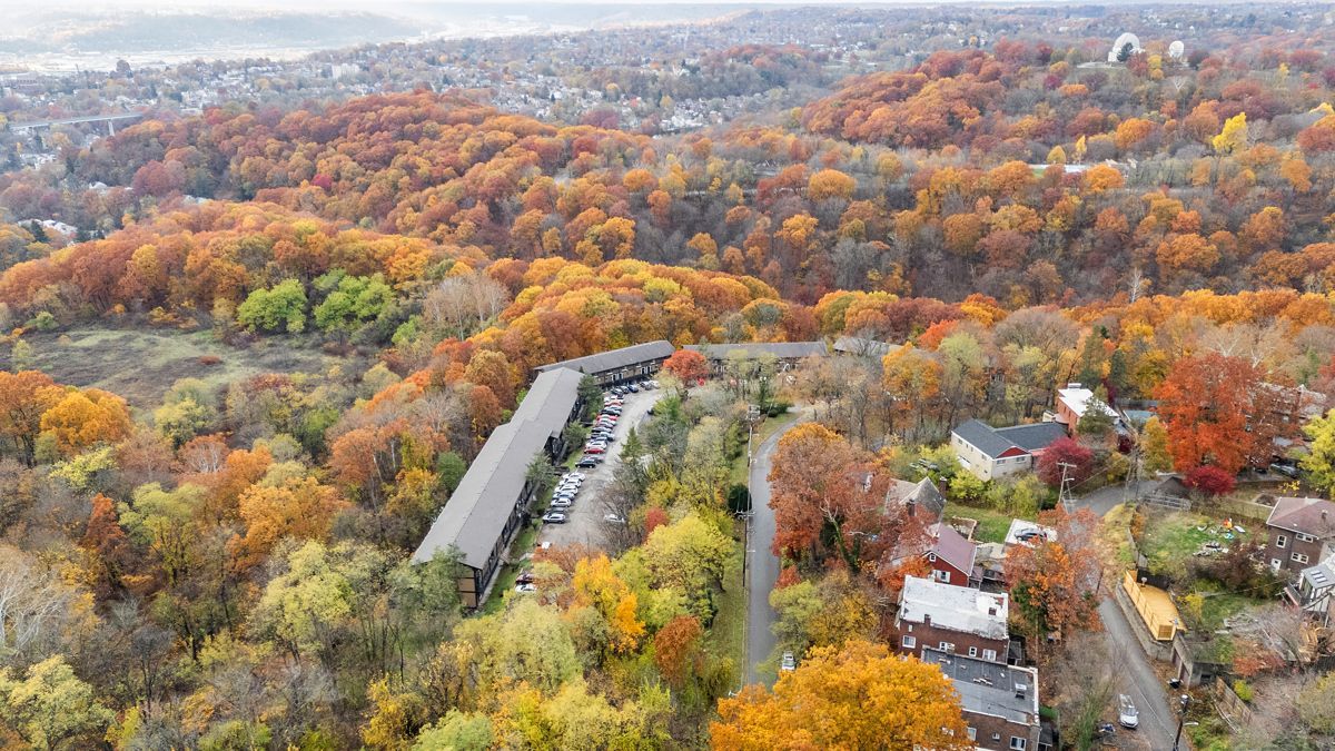 Autumnal view of a long apartment building and houses nestled amidst vibrant orange and yellow trees, hills, and a distant cityscape.