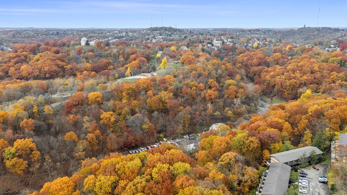 Vibrant autumn foliage covers a valley with a stream, buildings, and a distant town under a blue sky.