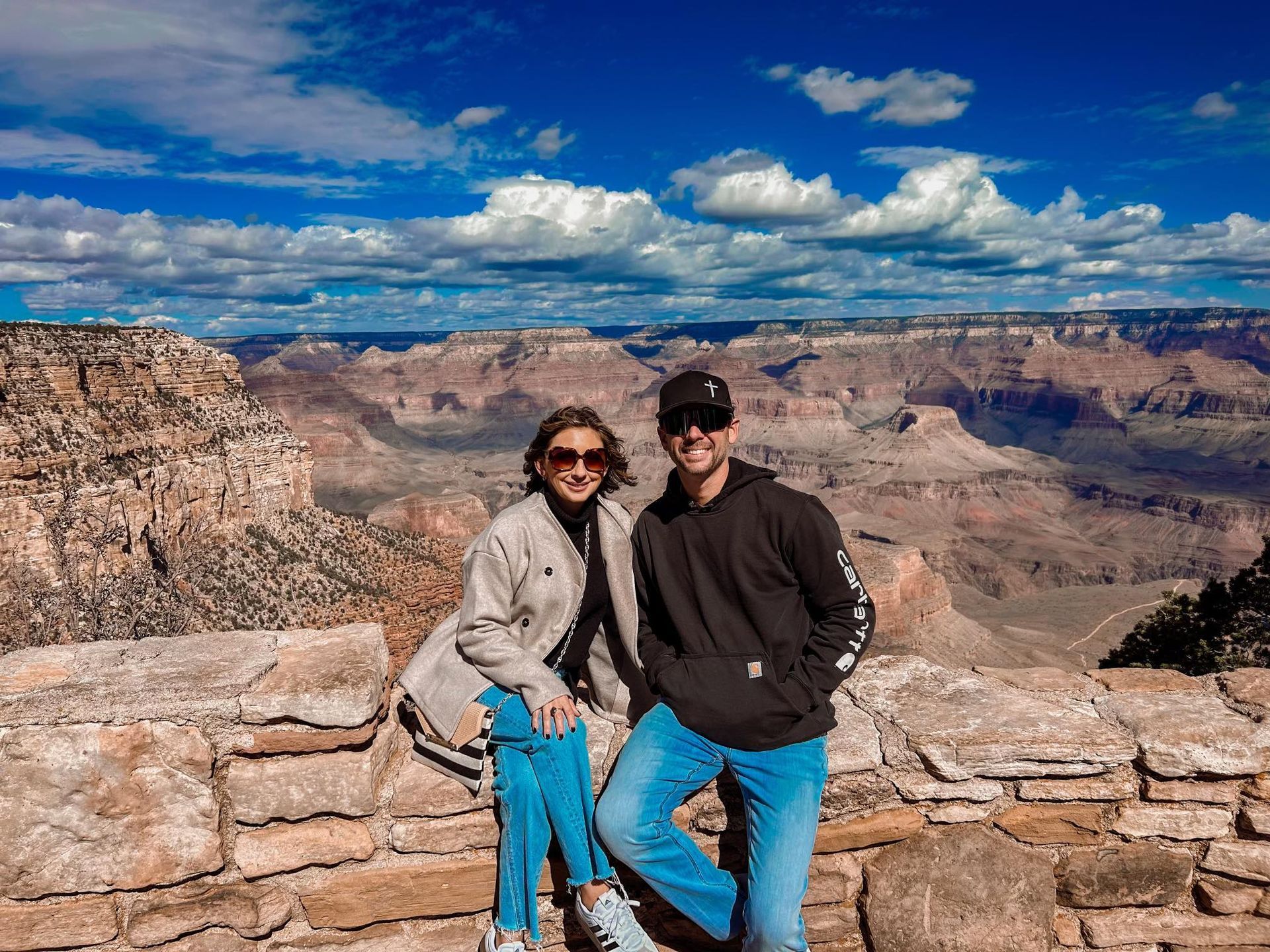 Couple poses on a stone wall overlooking the Grand Canyon. Blue sky with clouds.