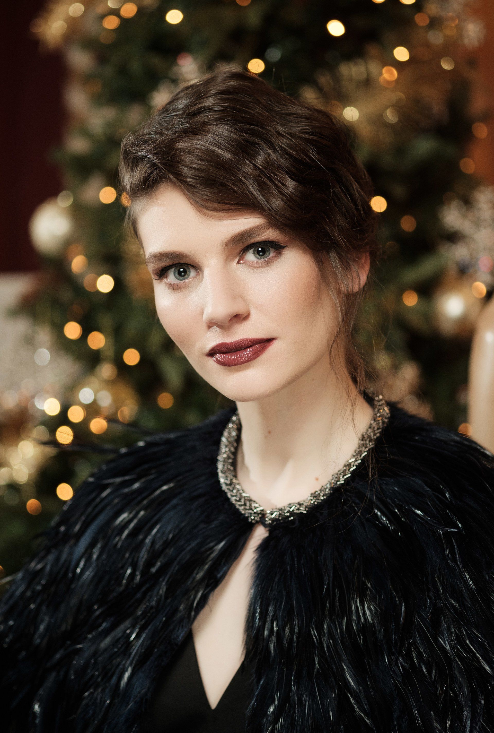 Pale woman with soft lips and eyes stood in front of Christmas tree looking at camera