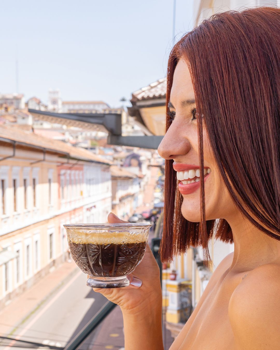 A smilig woman is holding a cup of coffee on a balcony.
