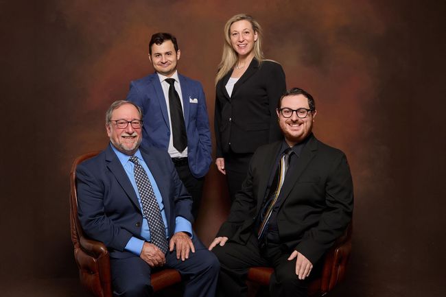 Four formally dressed people posing in a studio, two seated in chairs, against a brown backdrop.