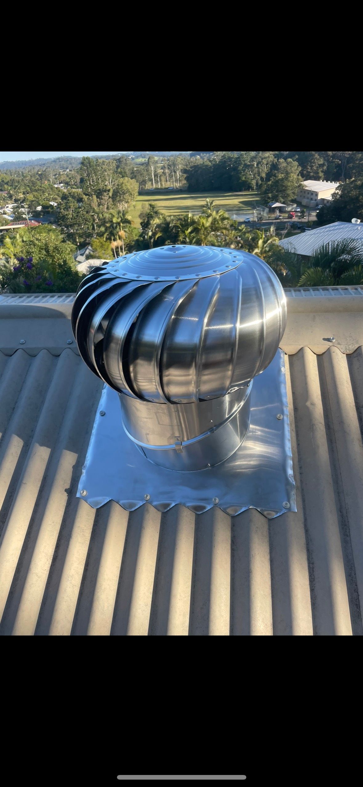 A shiny, stainless steel roof ventilator on a corrugated metal roof. In the background is greenery and a blue sky. — Whirly Bird King in Mango Hill, QLD