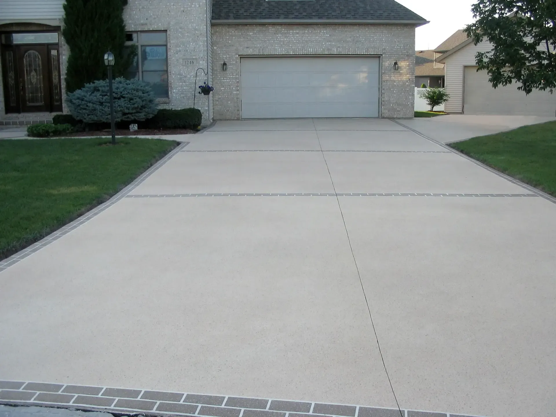 Driveway leading to a beige house with a garage, bordered by grass and decorative brick.