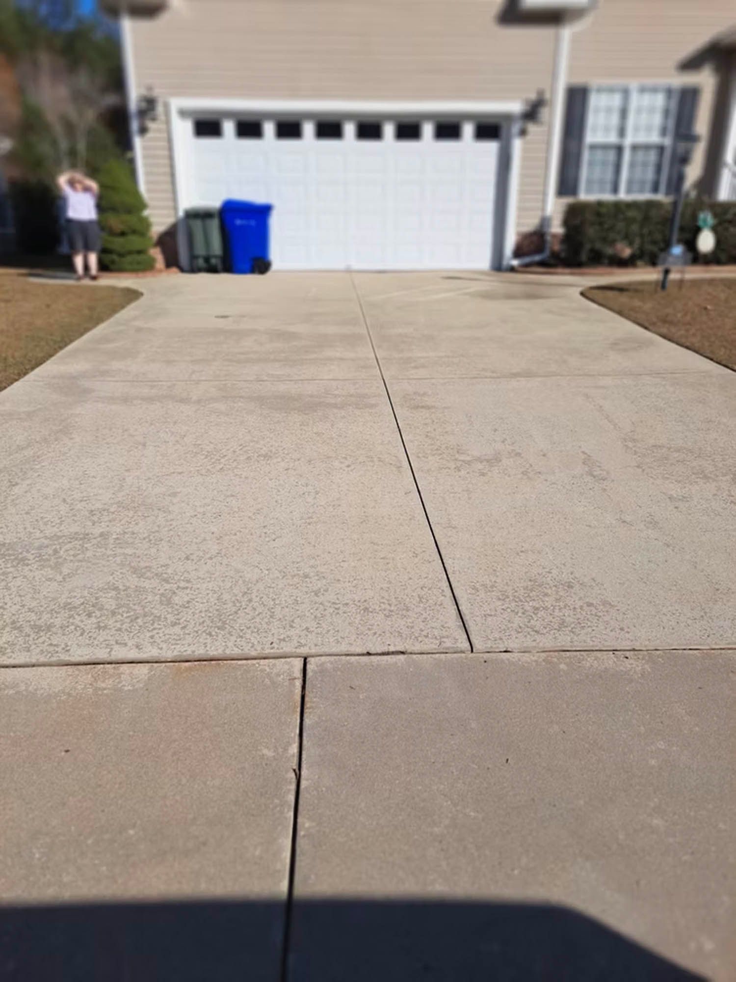 A concrete driveway leading to a house with a white garage door and a blue trash can.