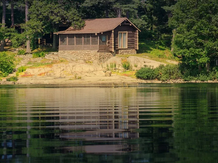 Log cabin by a lake, reflected in the water. Cabin has a brown roof and a screened-in porch, surrounded by trees.
