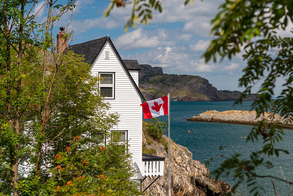 White house with Canadian flag on rocky coast overlooking ocean.