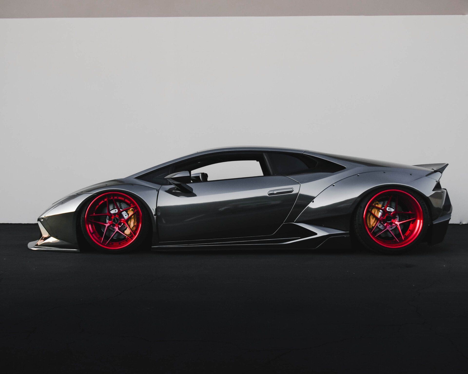 Gray Lamborghini with red rims parked against a white wall and dark ground.