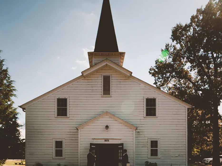 A white church with a steeple is surrounded by trees.