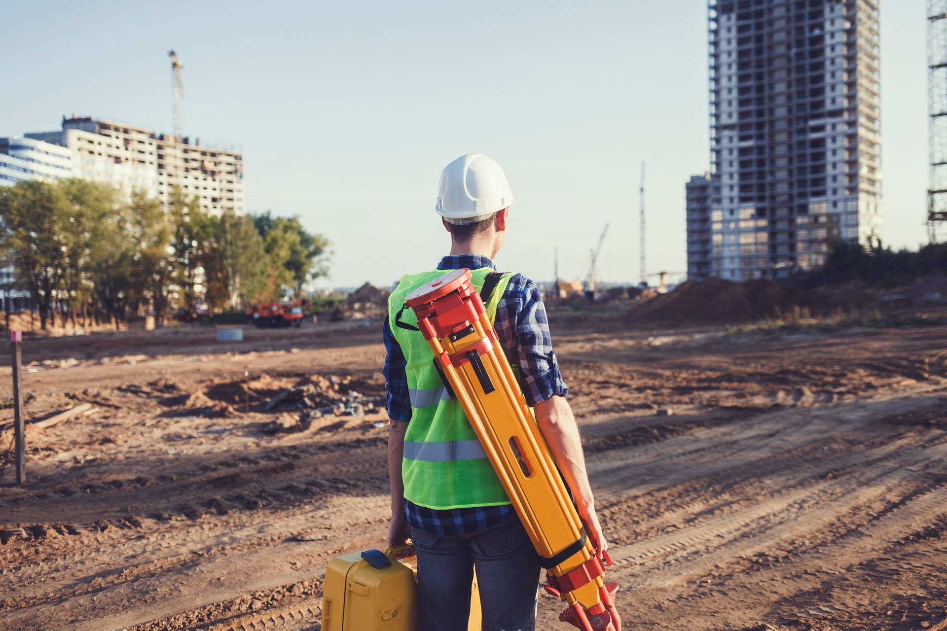 Un ouvrier du bâtiment, portant un casque et un gilet de sécurité, transporte du matériel de topographie et surveille un chantier.