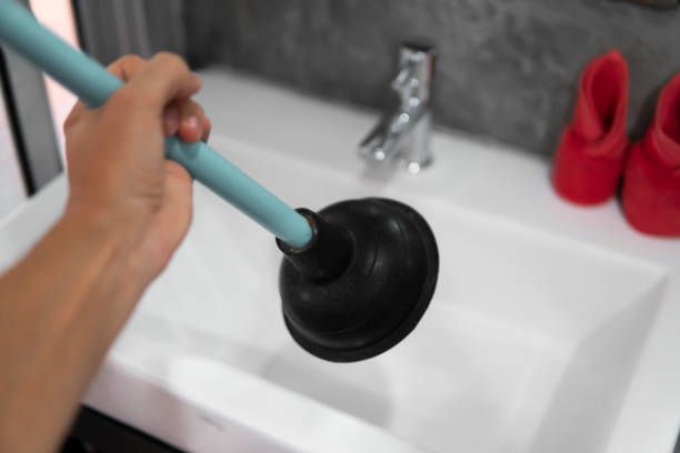 Person using a plunger in a white bathroom sink. A faucet is in the background, and red shoes are on the side.
