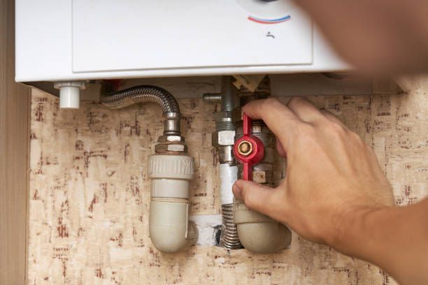 A hand turning a red valve on a white boiler, connected to pipes on a textured wall.