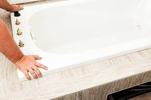 Person installing a white bathtub in a bathroom; hands visible, holding the tub.