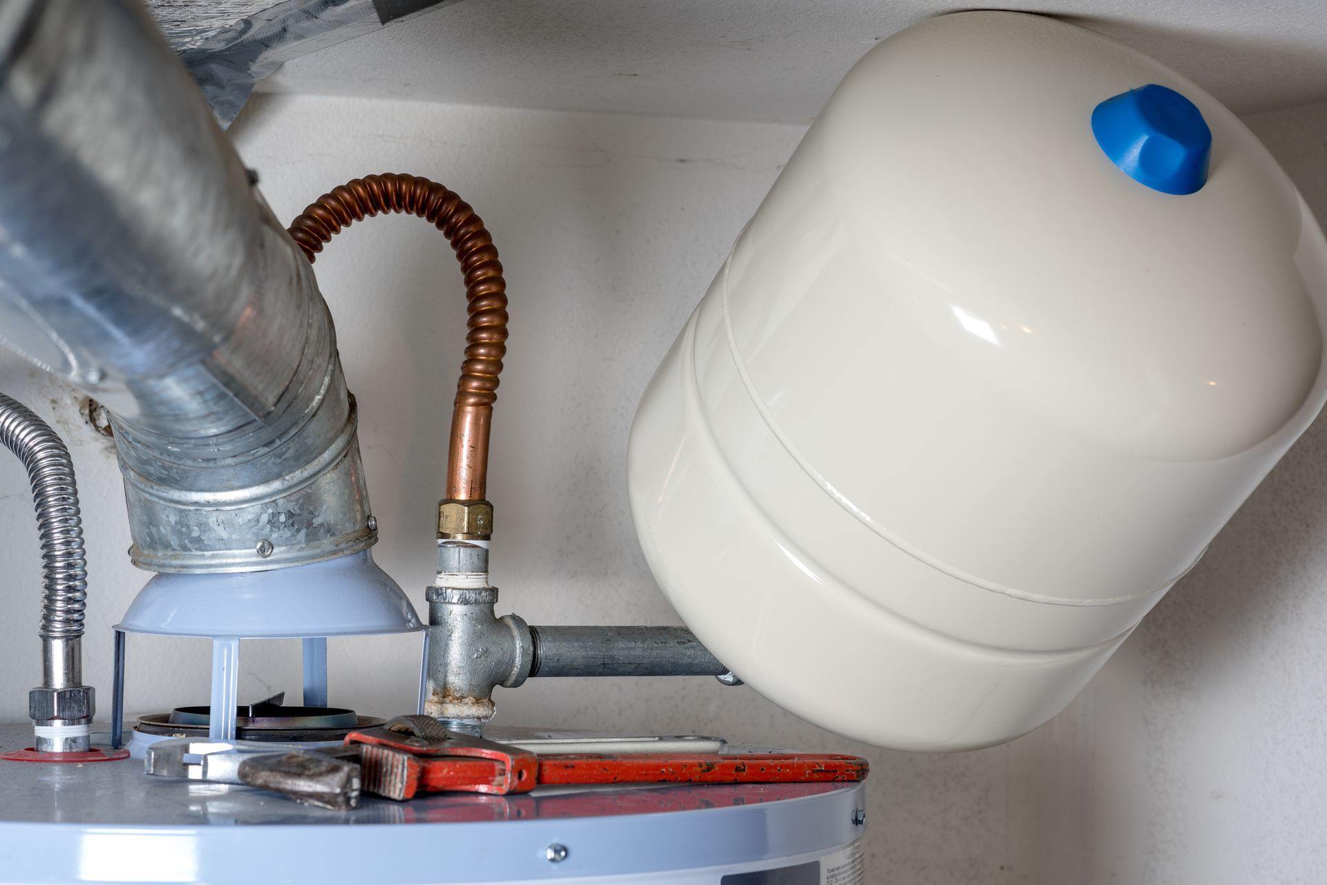 A white water heater tank with a blue top, connected to pipes and a pressure tank in a utility room.