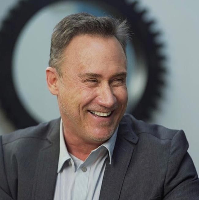 Smiling man in a gray blazer and light shirt in an office setting, with a circular black object behind him