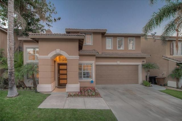 Two-story beige house with a lit front entry, garage, driveway, and small lawn at dusk