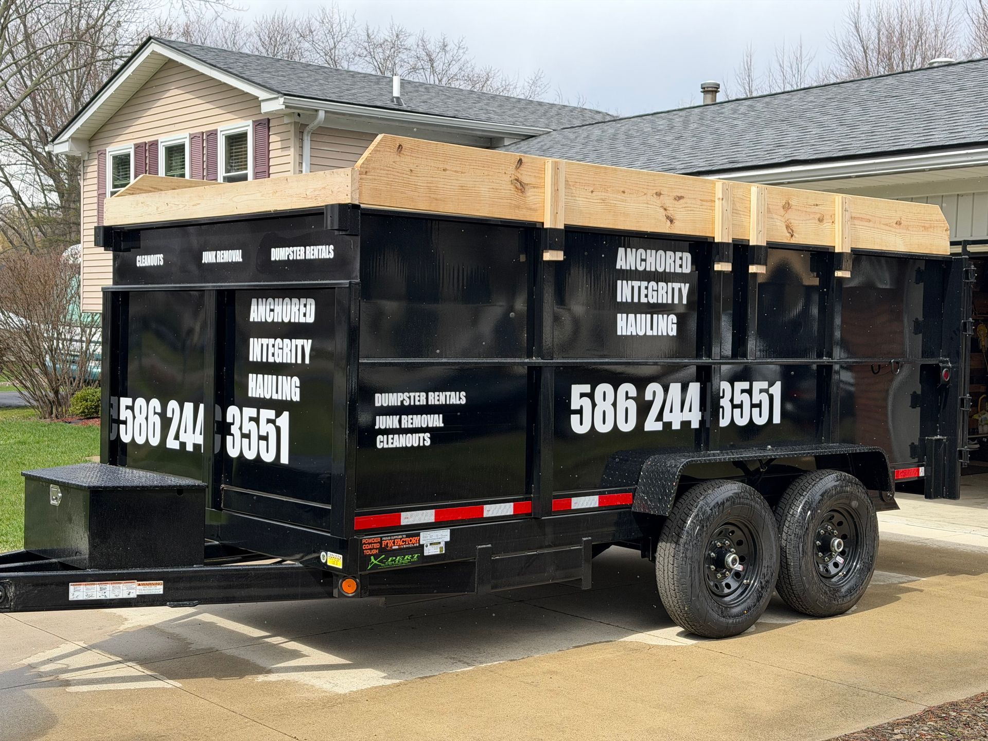 A black dump trailer parked on a concrete driveway in front of a house on a sunny day.