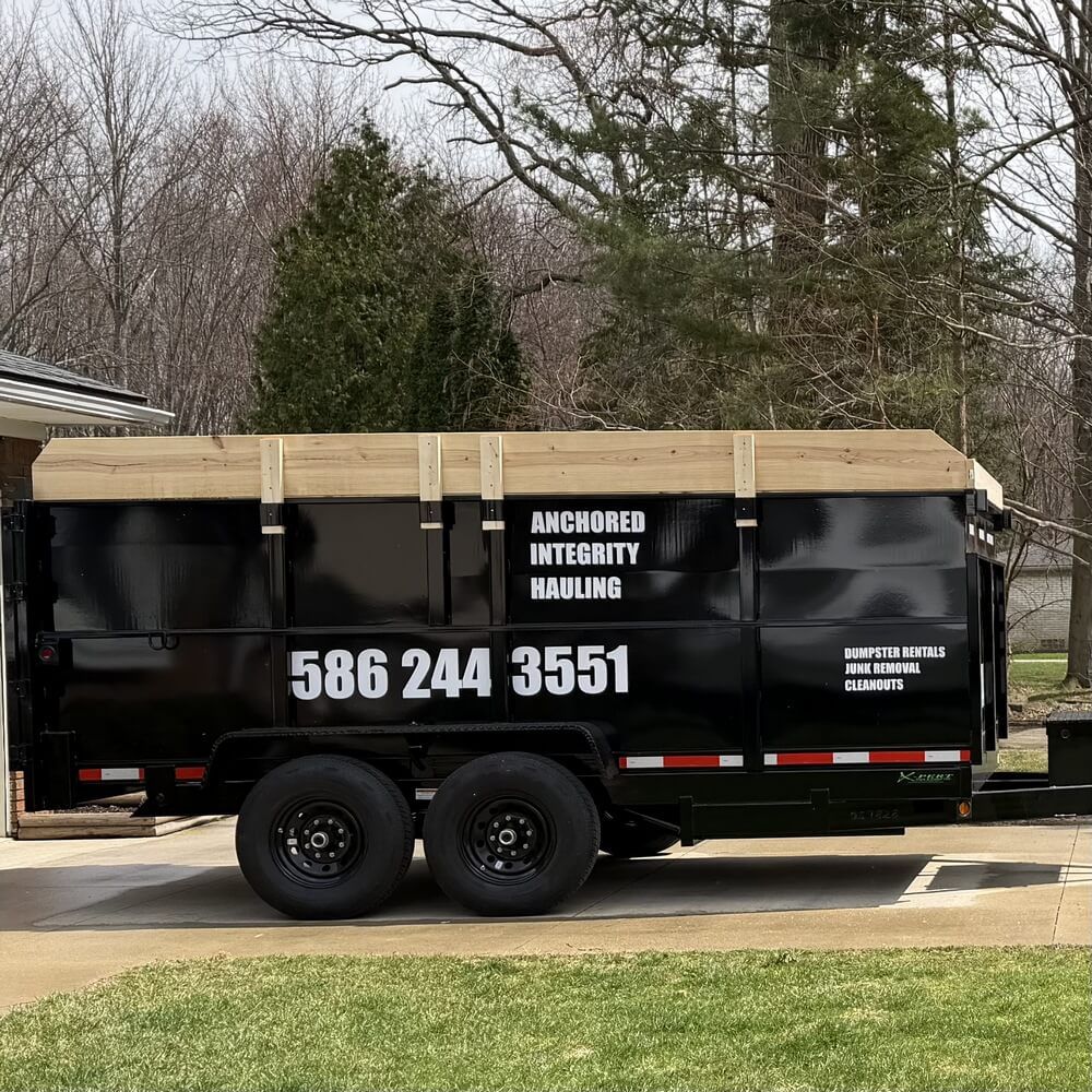 A black dump trailer parked on a concrete driveway in front of a house on a sunny day.