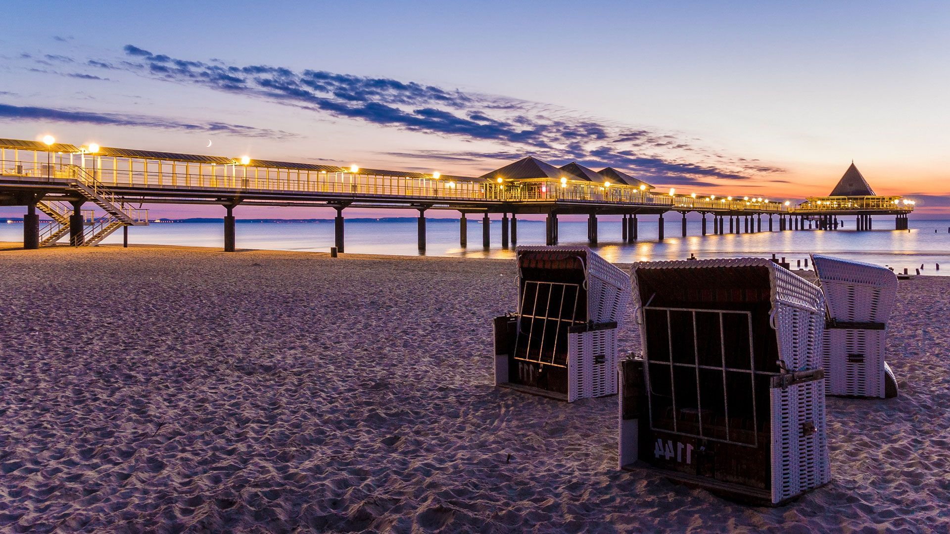 Une plage avec des chaises et une jetée en arrière-plan au coucher du soleil.