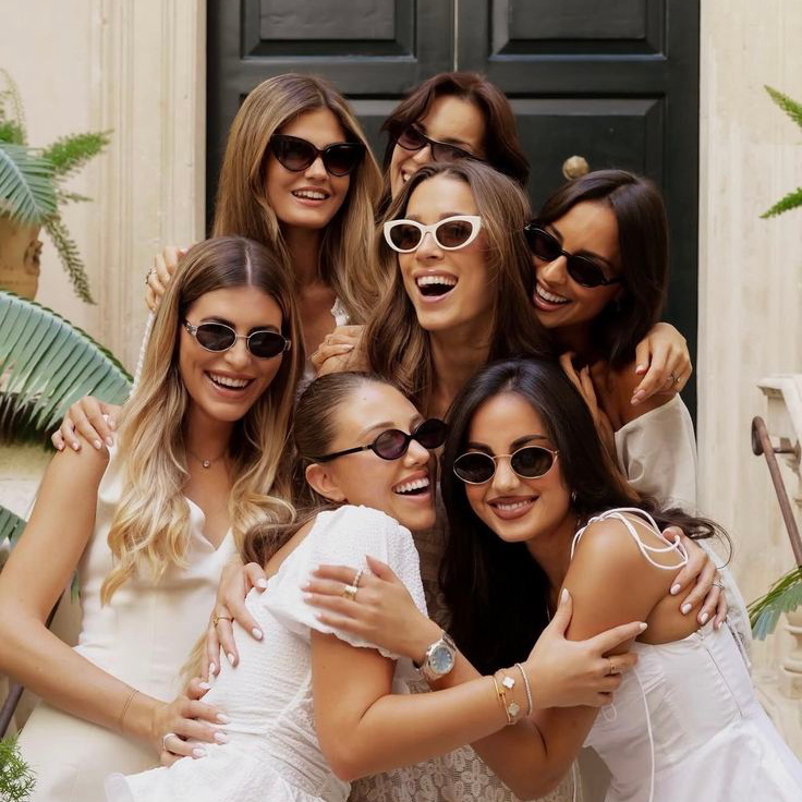 Six smiling women in white outfits and sunglasses pose together in front of a black door.