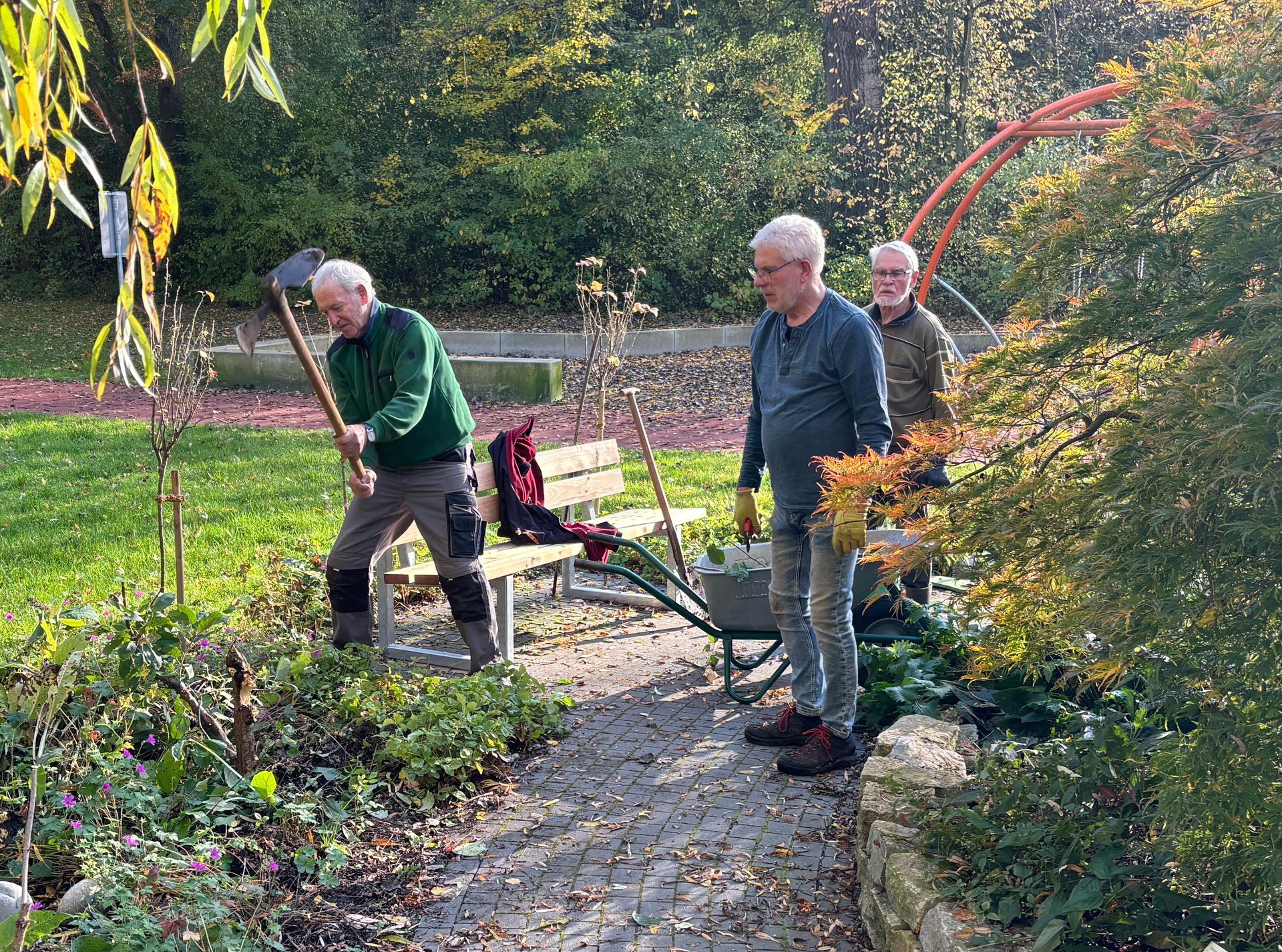 Drei Männer arbeiten im Freien im Garten; einer benutzt ein Gartengerät, zwei beobachten das Geschehen in der Nähe eines Gehwegs und einer Schubkarre.