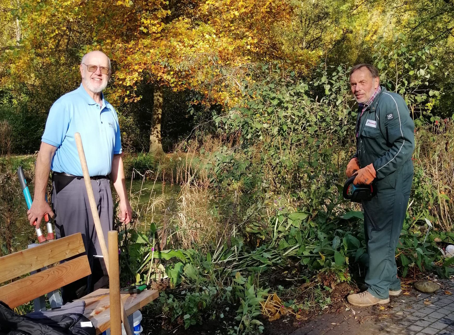 Zwei Männer neben wuchernden Pflanzen und einem Wasserlauf, einer in blauer, der andere in grüner Arbeitskleidung. Herbstlaub im Hintergrund.