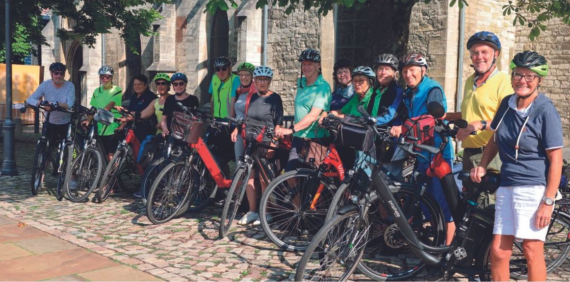 Eine Gruppe Radfahrer posiert mit ihren Fahrrädern vor einem Gebäude. Sie tragen Helme, farbenfrohe Kleidung und lächeln.