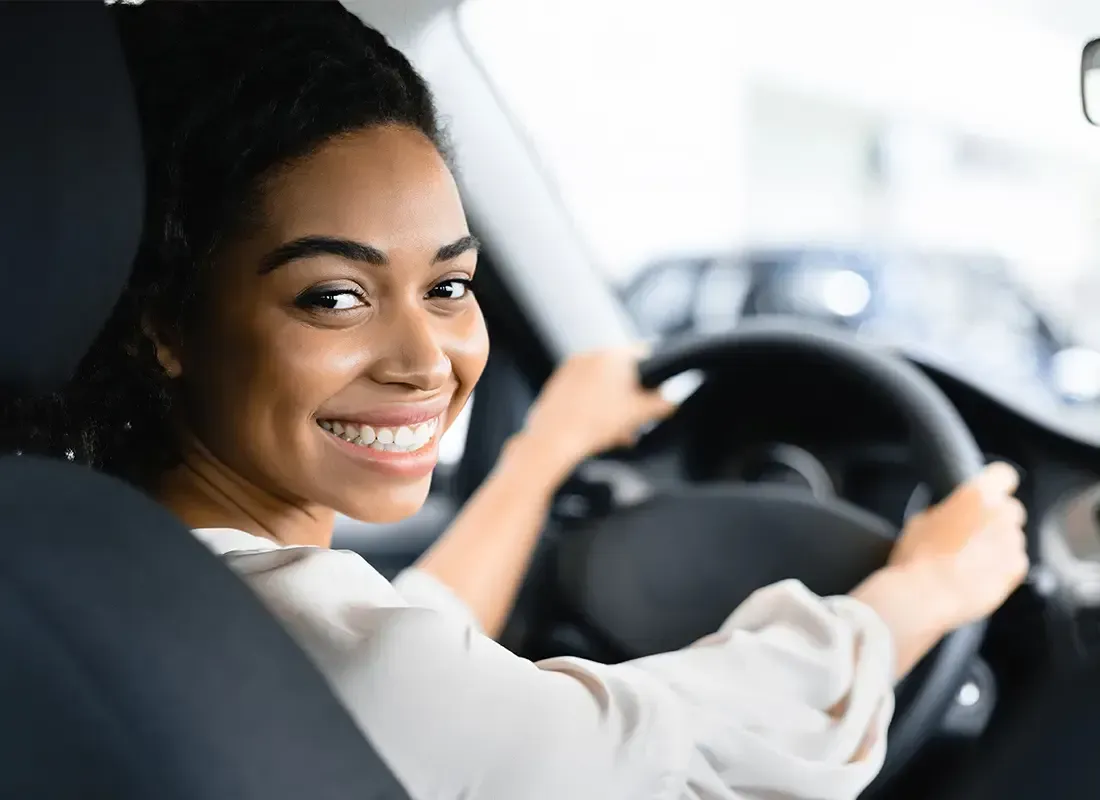 A smiling person sits in the driver's seat of a car, looking back toward the camera while holding the steering wheel.