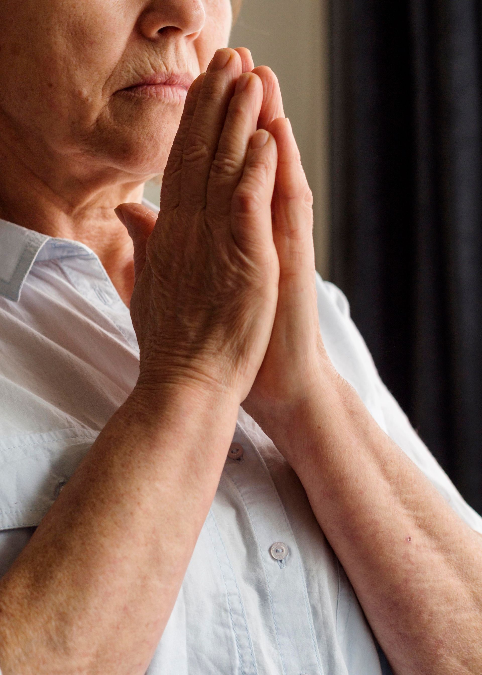 An elderly woman is praying with her hands folded in front of her face.