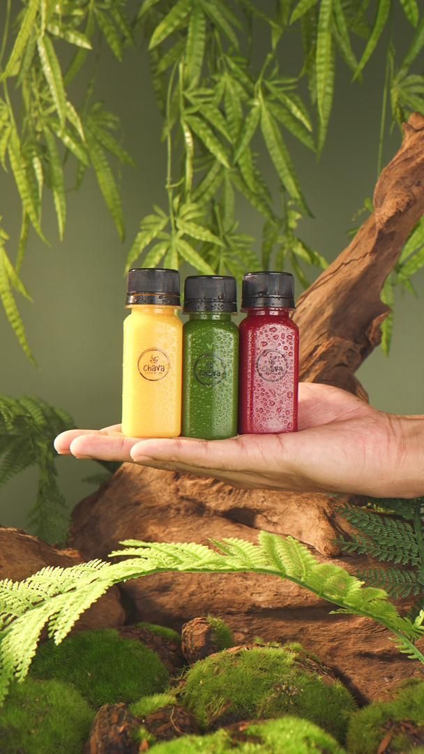 Hand holding three small juice bottles, yellow, green, and red, in a natural setting with foliage.