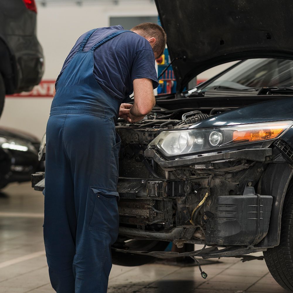 Mechanic in blue overalls working on a black car with an open hood in a garage.