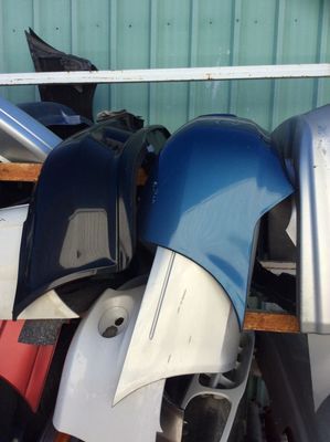 Pile of car bumpers in various colors, including blue, silver, and red, stacked outdoors.