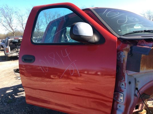 Red Ford truck door in a salvage yard with blue-tinted window and side mirror.